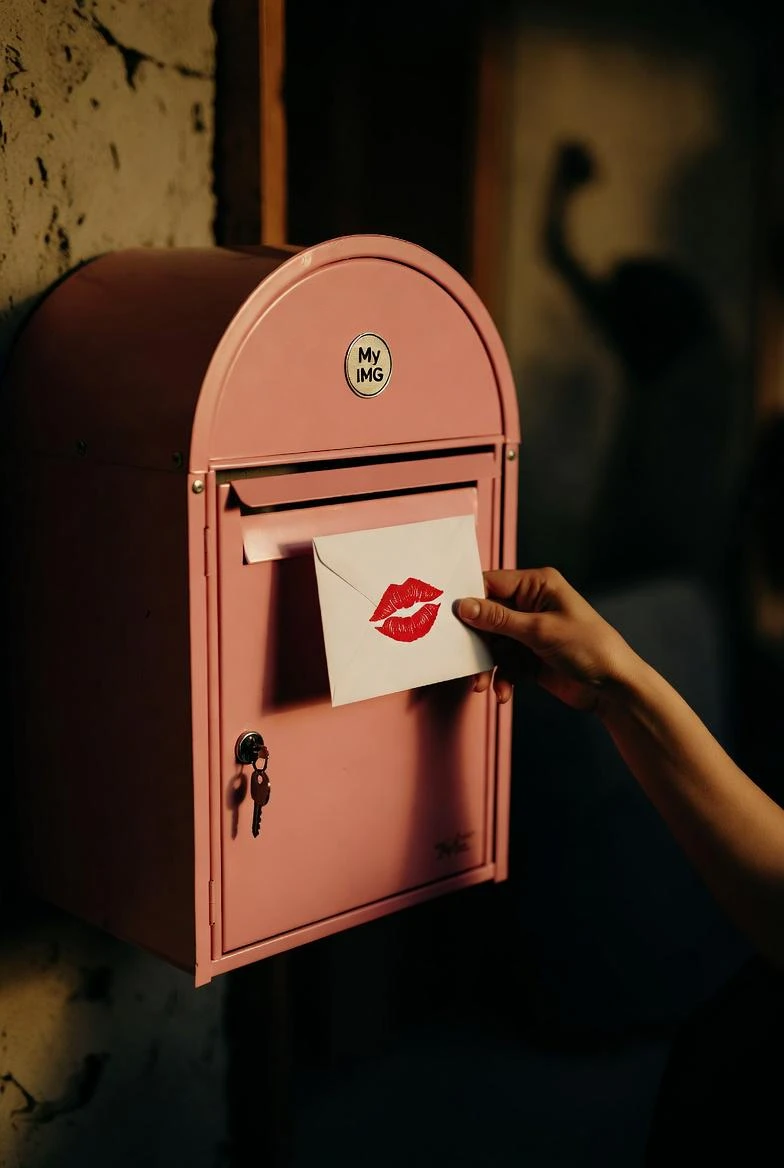 A hand placing a white envelope with a red lip print into a pink mailbox under warm, dim lighting.