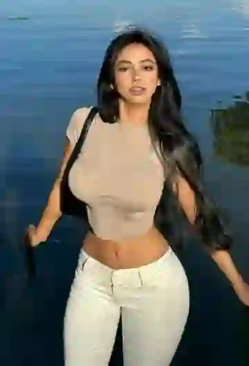 AI-generated image of a black-haired curvy woman wearing a gray crop top and white skinny jeans with a black shoulder bag, posing by a lake.