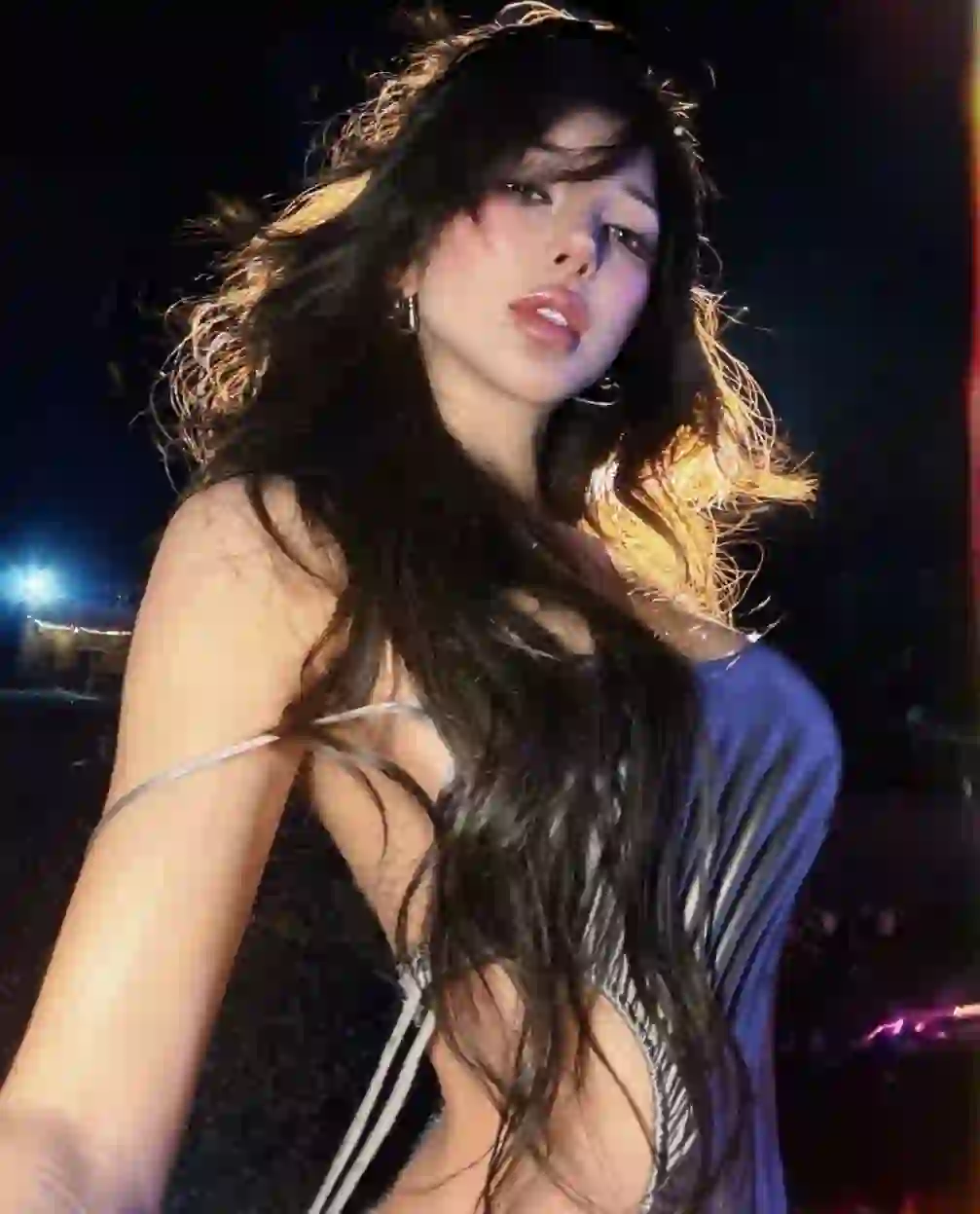 Low-angle shot of a woman with long black wavy hair wearing a sleeveless gradient grey dress, posing in a dimly lit space.