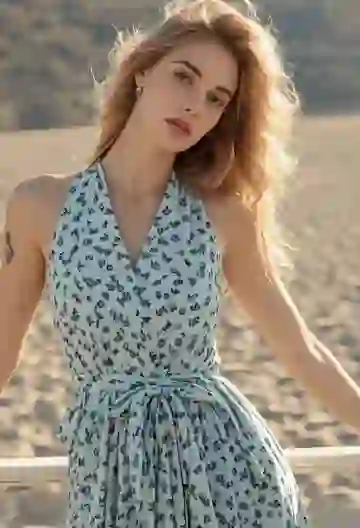 A blonde woman in a sleeveless blue floral dress stands against a white beach fence on a sunny day, tilting her head softly to the side.