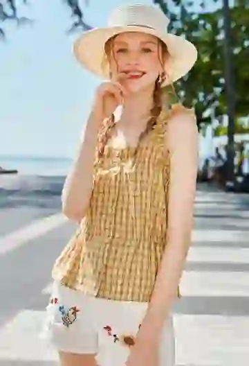 AI-generated image of a woman with two braids and a straw hat, wearing a brown-and-white plaid tank top and white cartoon-print shorts, posing playfully on a sunny street with her index finger near her mouth.