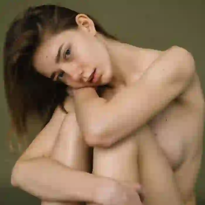 Portrait of a nude woman sitting with her legs drawn up to cover her body. Her right arm wraps around her legs, and her left arm rests on her knees.