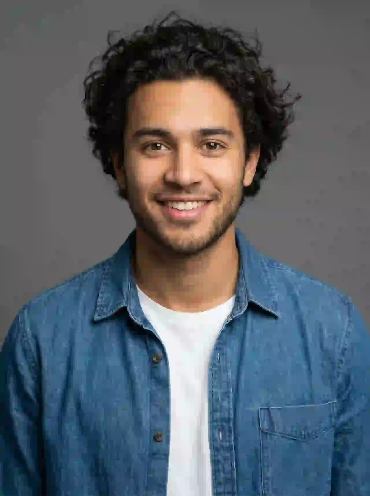 Young Middle Eastern man with curly hair in denim shirt smiling. Template for photo face swap.